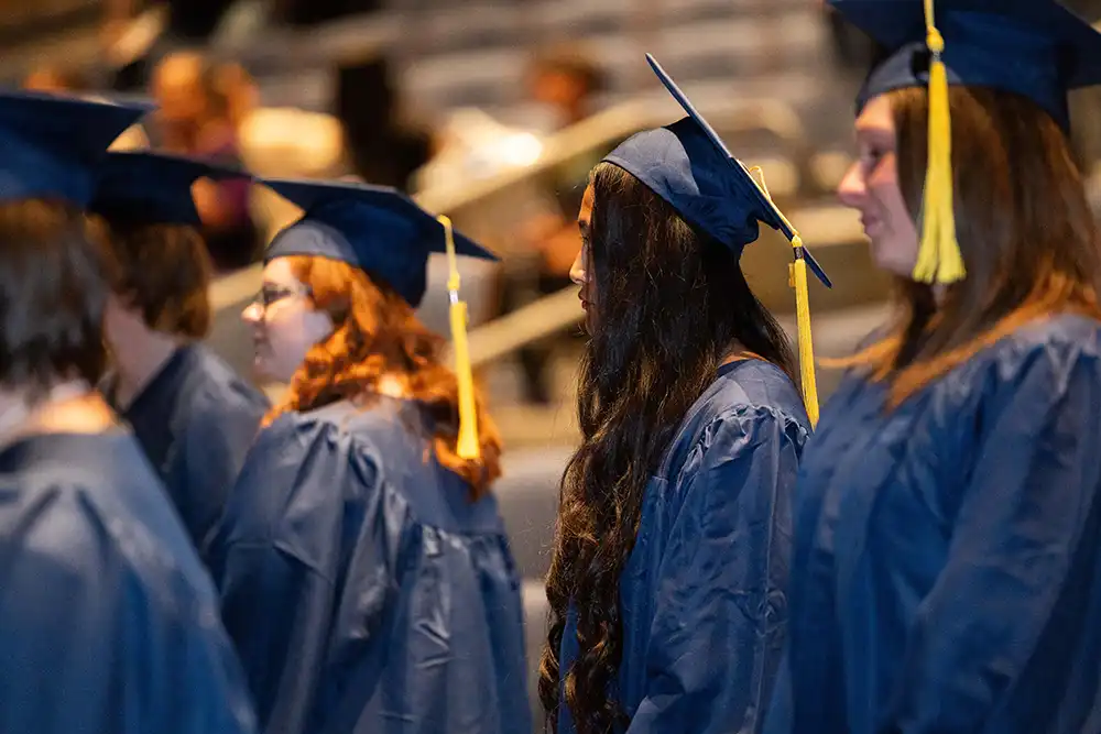 Students in blue graduation gowns and caps with gold tassels stand in a row during a commencement ceremony.